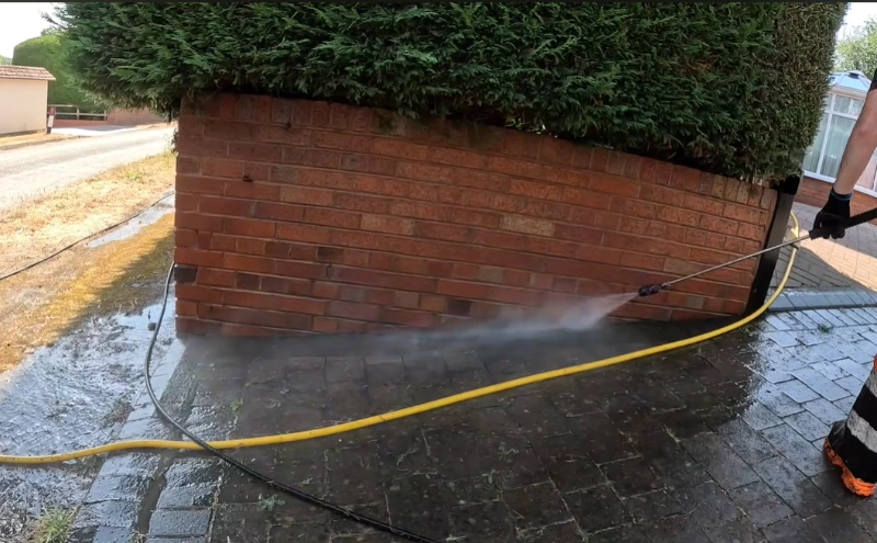 Technician rinsing a brick wall and driveway edge along a hedge-lined West Palm Beach property